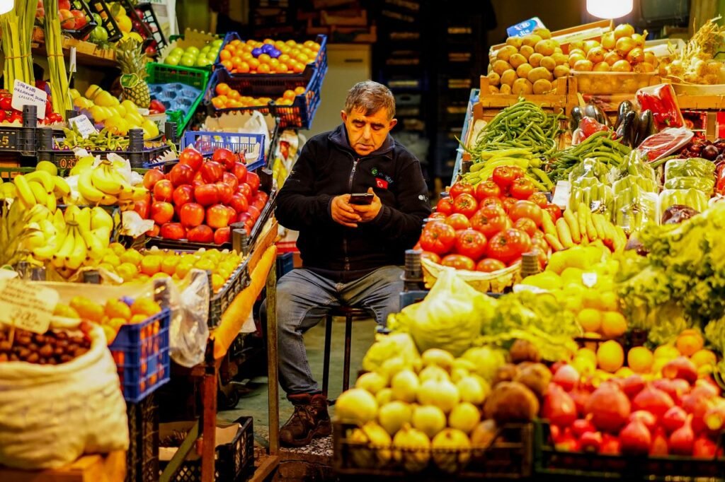 Man sitting in a colorful market stall, surrounded by fresh fruits and vegetables.